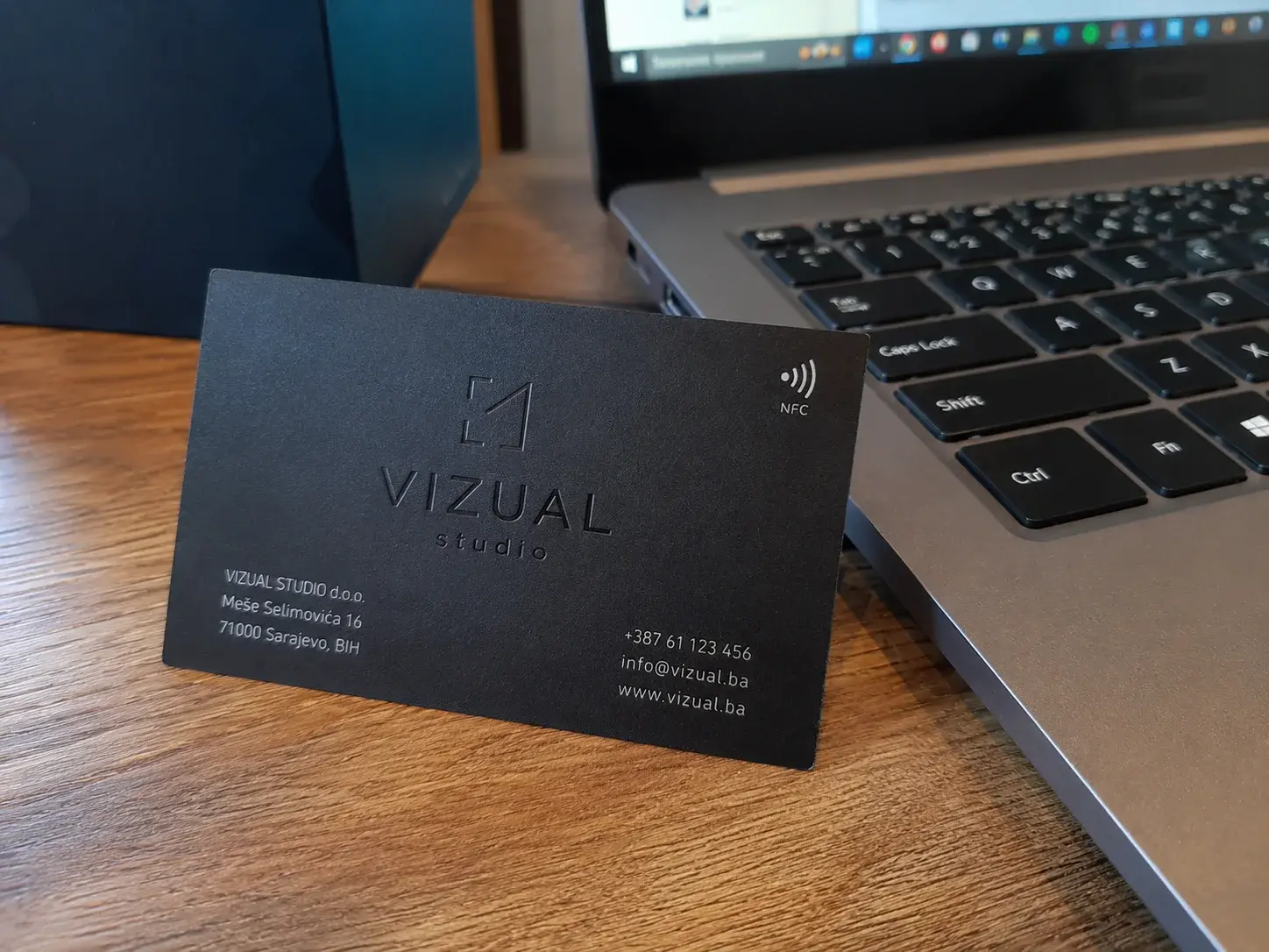 Crna mat NFC vizit kartica sa logo gravurom na stolu pored laptopa